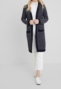 Marinblå lång kofta i strukturerad stickning, med diamantmönster, ribbade ärmslut, två franska fickor och öppen frontdesign.