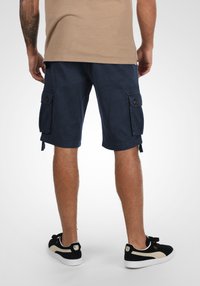 Marine Cargo-Shorts mit Seitentaschen, aus Baumwolle gefertigt. Mit einem Knopfverschluss und verstellbaren Säumen, getragen mit schwarzen Sneakers.