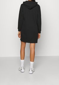 Robe à capuche noire avec manches longues, capuche réglable et coupe décontractée, associée à des chaussettes blanches et des chaussures de sport argentées.