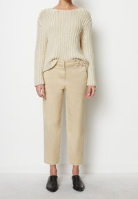 Persona che indossa un maglione a maglia grossa color crema, pantaloni beige a vita alta, e scarpe in pelle nere senza lacci, mentre è in piedi contro un muro bianco.