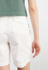 Personne portant un short écru et une chemise verte, vue de dos avec le bras droit détendu le long du corps.