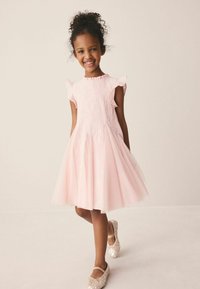 Lächendes Mädchen mit lockigem Haar, das ein hellrosa strukturiertes Kleid und glitzernde Ballerinas trägt, steht vor einem einfarbigen beigen Hintergrund.