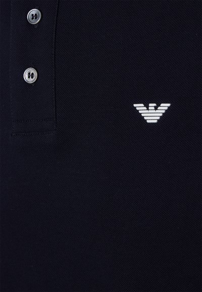 Marineblå polo skjorte med teksturert stoff, tre sølvknapper og en hvit, kantet logo på venstre bryst.