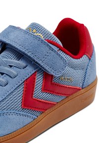 Niebieski zamszowy trampek z czerwonymi gumowymi akcentami, wyposażony w pasek na rzep, siateczkowy język oraz teksturowaną gumową podeszwę. Obecne złote logo.