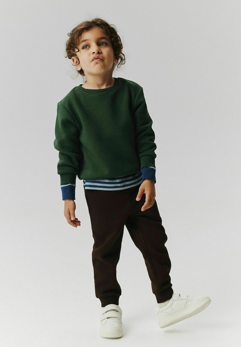 Enfant aux cheveux bouclés portant un sweat-shirt vert, un pantalon foncé et des baskets blanches, debout avec un pied légèrement soulevé.