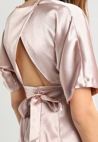Svetle růžové saténové šaty s krátkými nafouknutými rukávy, otevřeným designem na zádech a uvázanou mašlí v pase. Hladká textura a elegantní silueta.