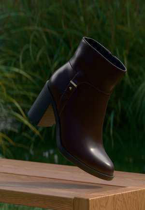 Bottine en cuir marron foncé avec talon épais et haut sur surface en bois, arrière-plan de feuillage vert flou.