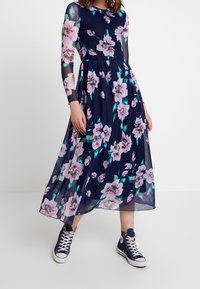 Robe fleurie à manches longues transparentes, base marine et fleurs roses. La jupe longue jusqu'aux chevilles est fluide, assortie à des baskets montantes navy.