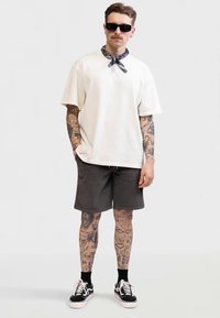 Mladic s tetovažama na rukama i nogama nosi bijelu oversized majicu, crne kratke hlače, crne tenisice, crne sunčane naočale i maramu oko vrata.
