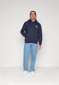 Sudadera azul marino con un diseño de bordado floral en el lado izquierdo del pecho, combinada con vaqueros de un azul claro y zapatillas blancas. Textura suave, ajuste casual.