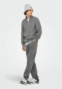 Grauer Zip-Kragen-Pullover aus weichem Material, kombiniert mit verblassten grauen Jeans und weißen Sneakers mit schwarzen Akzenten. Eine graue Mütze rundet das Outfit ab.