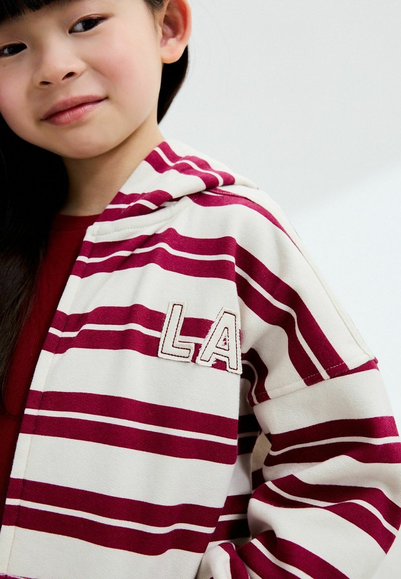 Sweatshirt à capuche en crème avec des rayures bordeaux, présentant un patch texturé "LA". Tissu doux avec une coupe décontractée, mettant en avant la capuche proéminente.