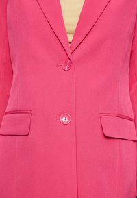 Blazer rose vif à texture douce, avec deux poches avant et deux boutons ronds. Coupe ajustée avec épaules structurées grâce aux épaulettes.