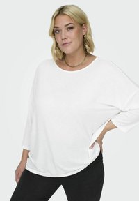 Top blanco de gran tamaño con mangas largas, cuello ancho y ajuste holgado, combinado con pantalones negros ajustados. Textura suave y diseño minimalista.