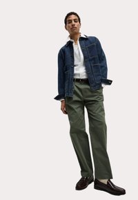 Chaqueta de mezclilla con bolsillos frontales, camisa blanca de cuello, pantalones verdes y zapatos marrón oscuro. Outfit casual en capas con un ajuste relajado.