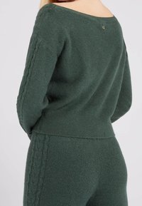 Femme portant un pull en maille texturée vert foncé et un pantalon assorti, vue de dos et de profil, sur un fond clair uni.
