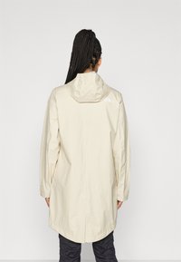 Veste imperméable ample de couleur crème avec capuche, manches longues et ourlet arrondi. Le logo est placé près du col.