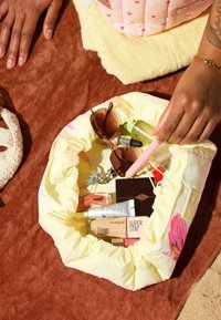 Des mains plongent dans une pochette matelassée jaune contenant des lunettes de soleil, des articles de maquillage, un recourbe-cils et des produits de soin de la peau sur une surface marron.
