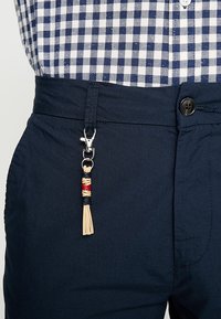 Camicia a quadri blu e bianchi infilata in pantaloni blu navy, con un piccolo ciondolo a nappina beige, rosso e blu attaccato a un passante della cintura.
