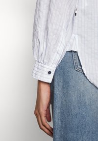 Personne portant une chemise blanche à manches longues avec des fines rayures bleues et un jean bleu en denim, montrant le poignet avec un bouton noir et une main détendue.