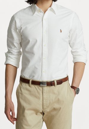 Mann, der ein weißes Hemd mit Knöpfen und kleinem gesticktem Logo, beige Hosen, braunen Ledergürtel und Armbanduhr trägt, steht mit einer Hand in der Tasche.