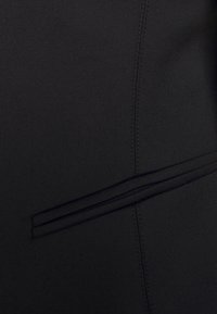 Gros plan sur un tissu noir avec une poche passepoilée horizontale et un détail de couture verticale visible.