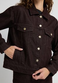 Giacca in denim marrone scuro con un design corto, bottoni a pressione dorati, due tasche sul petto e tasche laterali. Tessuto testurizzato con cuciture visibili.