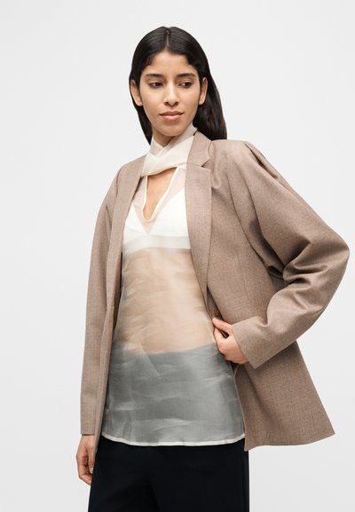 Blazer marron sur mesure avec épaules structurées, superposé à un haut crème semi-transparent avec un ourlet gris. Textures lisses et lignes épurées.