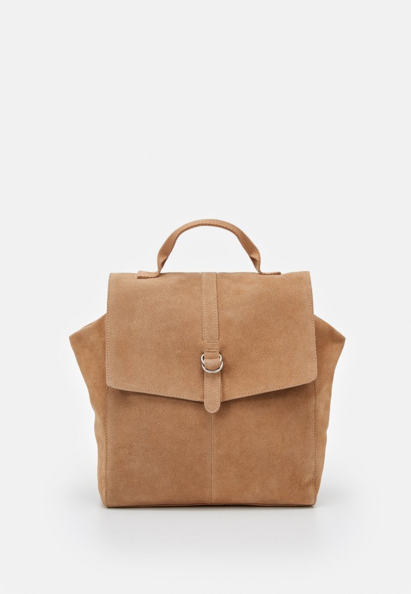 Sac à dos en daim beige avec poignée supérieure et rabat avant sécurisé par une lanière avec une boucle argentée, présenté sur un fond clair uni.