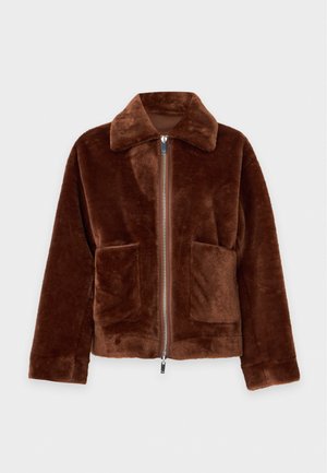 Un mannequin se tient devant un fond blanc uni, vêtu d’une veste marron en fausse fourrure, d’un haut marron, d’un pantalon crème et de baskets beiges.