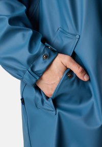 Wodoodporna niebieska kurtka przeciwdeszczowa z guzikami przy mankietach, kieszenią z przodu oraz gładkim, teksturowanym materiałem.