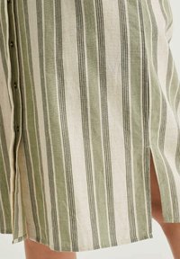 Robe en lin rayée verte et beige, présentant des lignes verticales, des détails de boutons et des fentes latérales pour plus de liberté de mouvement.