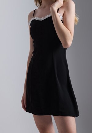 Femme portant une robe noire sans manches avec un bord volanté blanc au niveau de l'encolure, debout devant un fond gris.