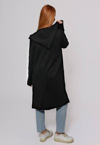 Cappotto lungo nero con cappuccio, realizzato in un tessuto testurizzato. Presenta maniche ampie e tasche laterali, indossato sopra jeans azzurri chiari e sneaker bianche.