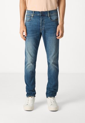 Blaue Jeans aus Denim mit geradem Schnitt, Fronttaschen und subtilen Abnutzungserscheinungen. Kombiniert mit weißen Sneakers.