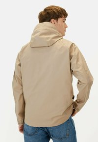 Beige letvægtsjakke med hætte, der har lange ærmer og en glat tekstur. Bemærkelsesværdige detaljer inkluderer diskret branding på siden.