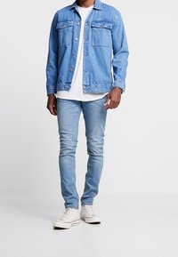 Světle modrá džínová bunda se dvěma kapsami na hrudi, nošená přes bílé tričko, v kombinaci se světle modrými úzkými džíny a bílými teniskami.