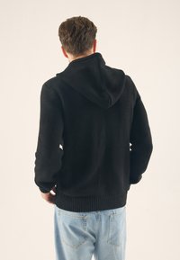 Svart stickad huvtröja med en texturerad tyg, ribbade muddar och fåll. Har en rymlig huva och bärs över ljusblå jeans.