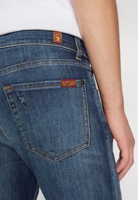 Jean en denim bleu foncé avec une poche arrière déchirée, comportant une étiquette rouge avec du texte jaune et des détails de couture traditionnels.