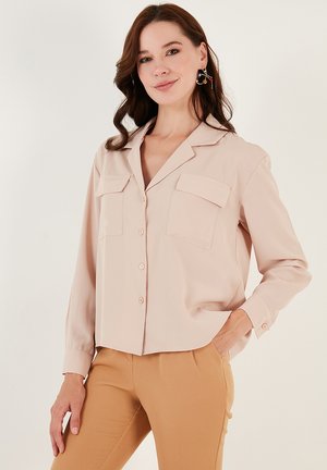 Light pink blouse with long sleeves, button-up front, two chest pockets, and a soft, smooth fabric texture. Paired with beige pants.