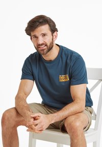 Man with dark hair and beard wearing blue "Endless Summer" t-shirt and khaki shorts, sitting on white chair with hands clasped.