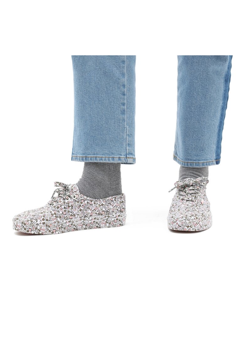 Chaussures à lacets à motif floral en tissu gris clair avec une surface texturée, associées à des chaussettes de cheville grises et un pantalon en denim bleu.