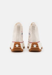 Beżowe sneakersy typu high-top z materiału kanvas, z naszywką logo na pięcie, gumową podeszwą z detalem w kształcie gwiazdki oraz kontrastową brązową podeszwą.