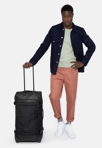 Borsa da viaggio nera con rotelle, realizzata in tessuto resistente, di forma rettangolare, con tasche laterali. Il modello indossa una giacca scura, una camicia chiara e pantaloni rosa.