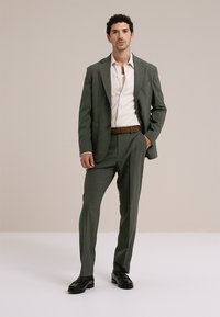 Pilkas pritaikytas kostiumas su tekstūriniu paviršiumi, sudarytas iš švarko su įpjauta atlape ir derančių kelnių. Dėvimas ant balto marškinio, rudos spalvos diržo ir tamsių batų.