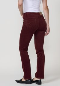 Vrouw in nauwsluitende donkerrode corduroy broek en zwarte instappers, staande met haar rug naar de camera tegen een effen achtergrond.