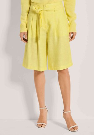 Shorts en lin jaune avec une taille ceinturée, présentant des plis sur le devant et une coupe ample, assortis à des talons nus à lanières.