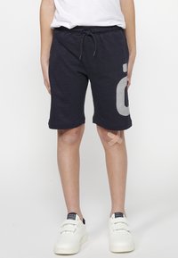 Marineblaue Baumwollshorts mit elastischem Bund, Kordelzug und weißen gestreiften grafischen Details am rechten Bein. Getragen mit weißen Sneakern.