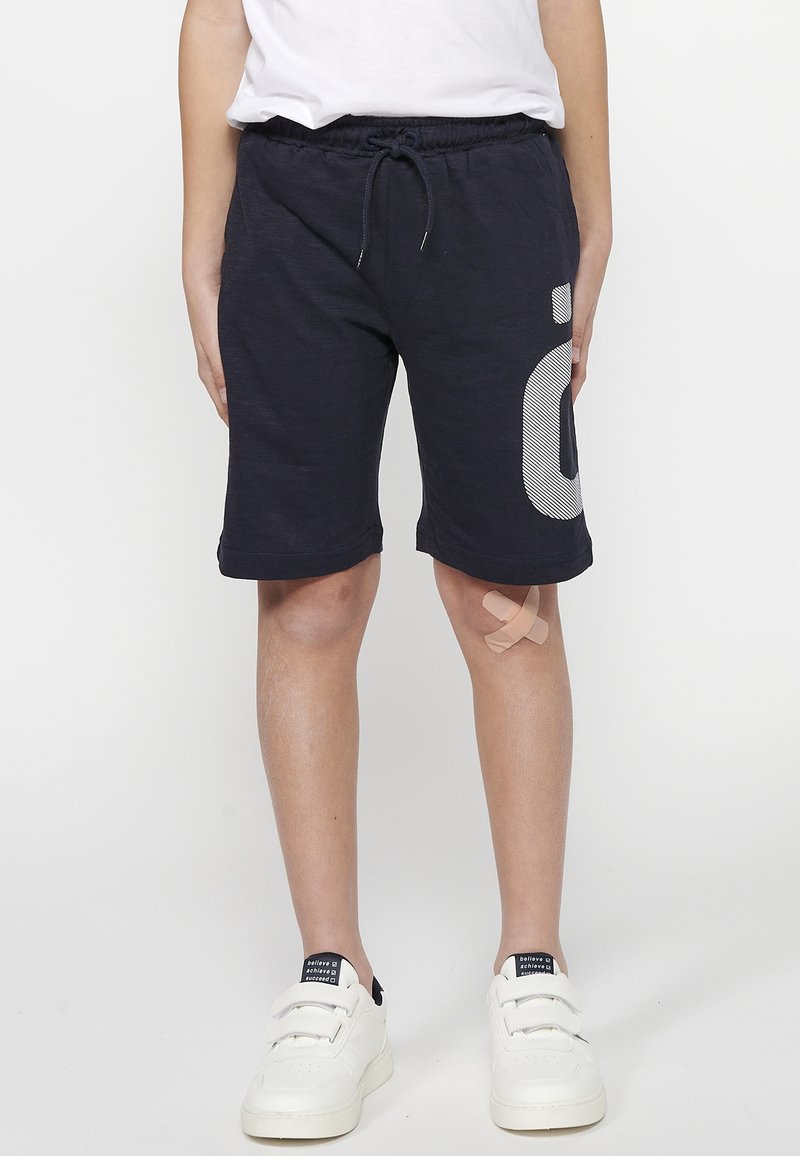 Marineblaue Baumwollshorts mit elastischem Bund, Kordelzug und weißen gestreiften grafischen Details am rechten Bein. Getragen mit weißen Sneakern.