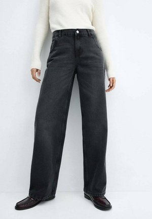 Persoon die hoge taille, wijde zwarte jeans draagt, een witte trui met lange mouwen en donkerbruine loafers, staand tegen een witte achtergrond.